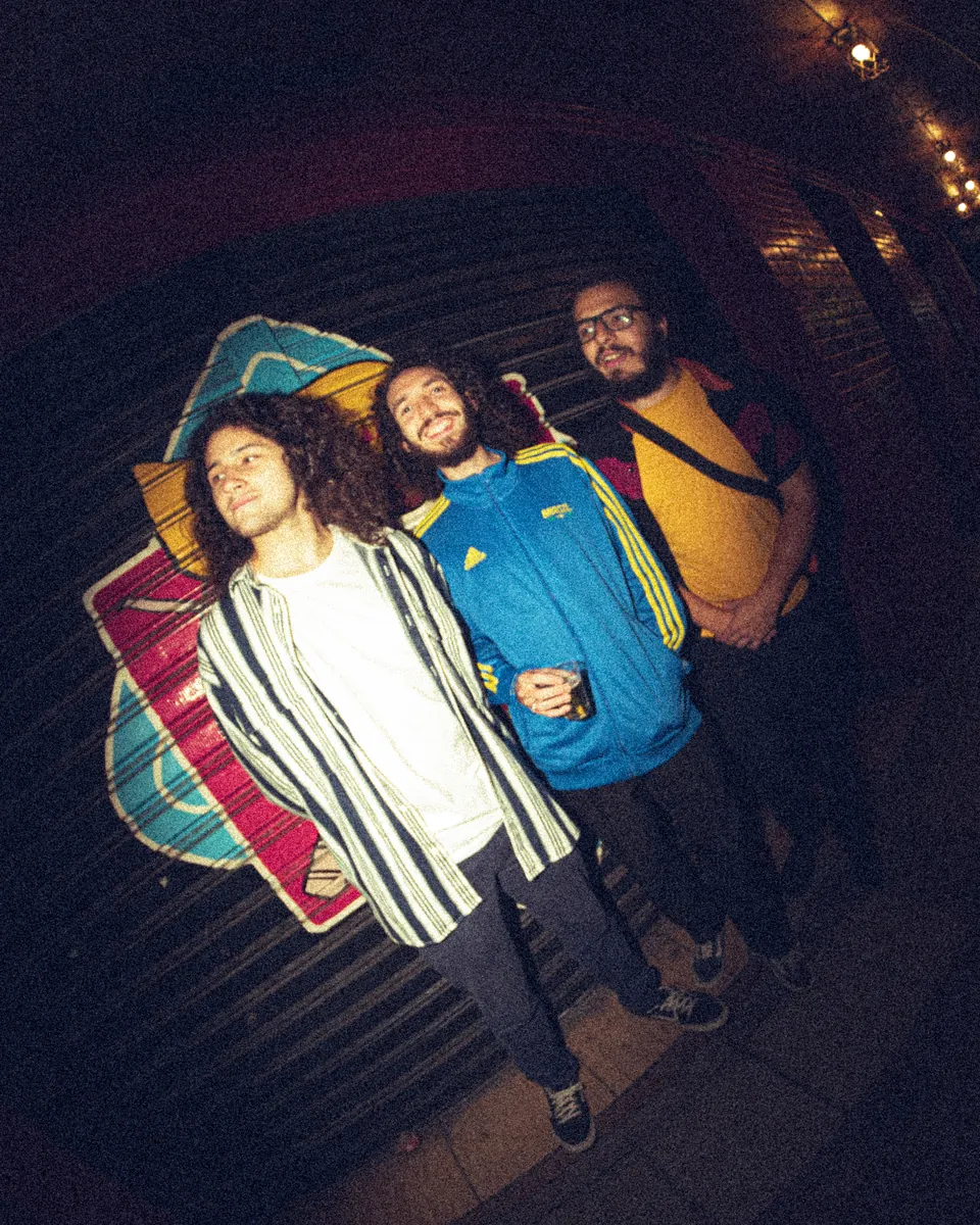 Os três músicos juntos em um salão de sinuca, fotografados com lente grande angular.