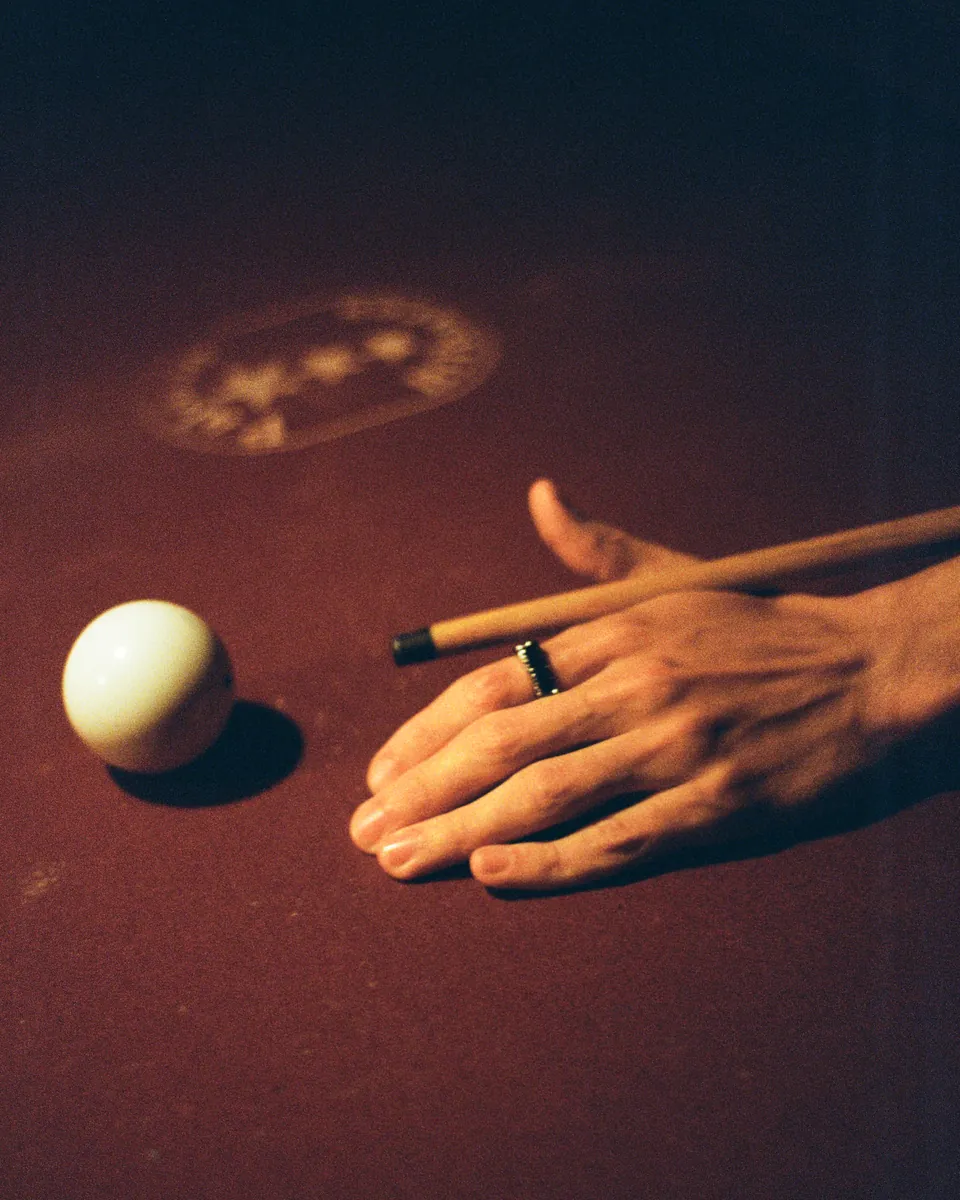 Mão e taco próximos a uma bola de sinuca, em fotografia analógica.