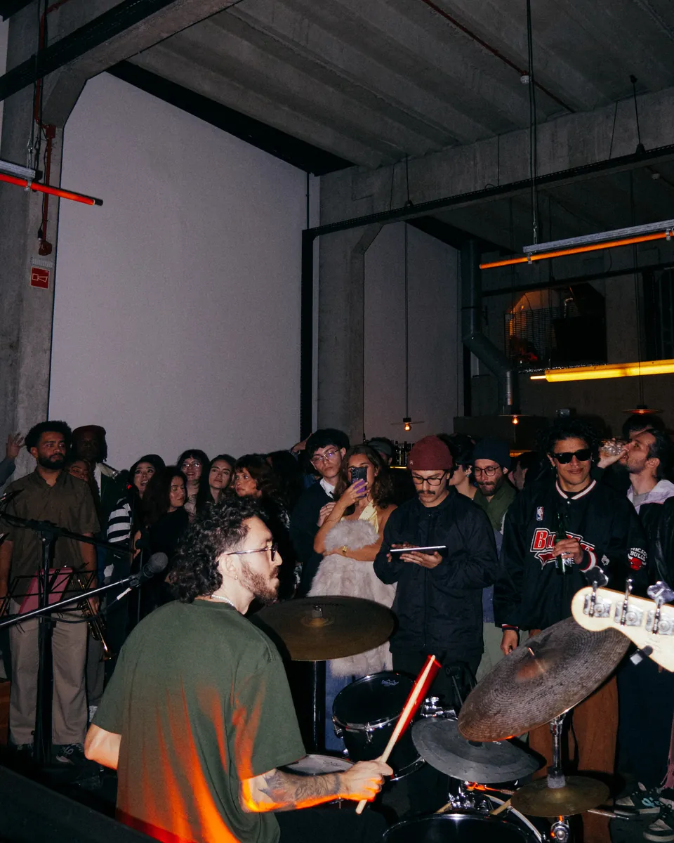 Bateria vista por trás, com músicos e público em uma sala próxima.