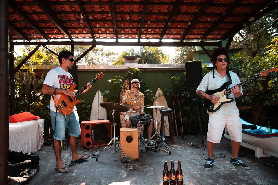 Los Mudos tocando baixo, bateria e guitarra sob um telhado de madeira.